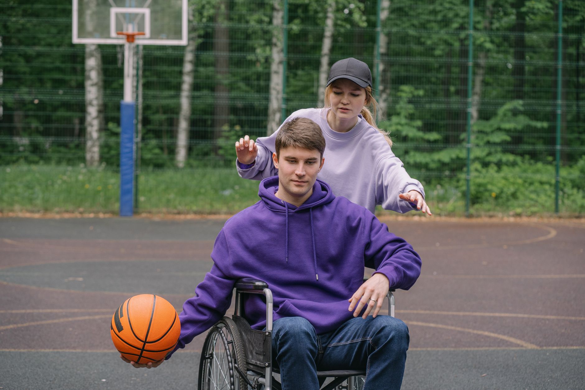 A woman assists a man in a wheelchair playing basketball outdoors, promoting inclusivity.