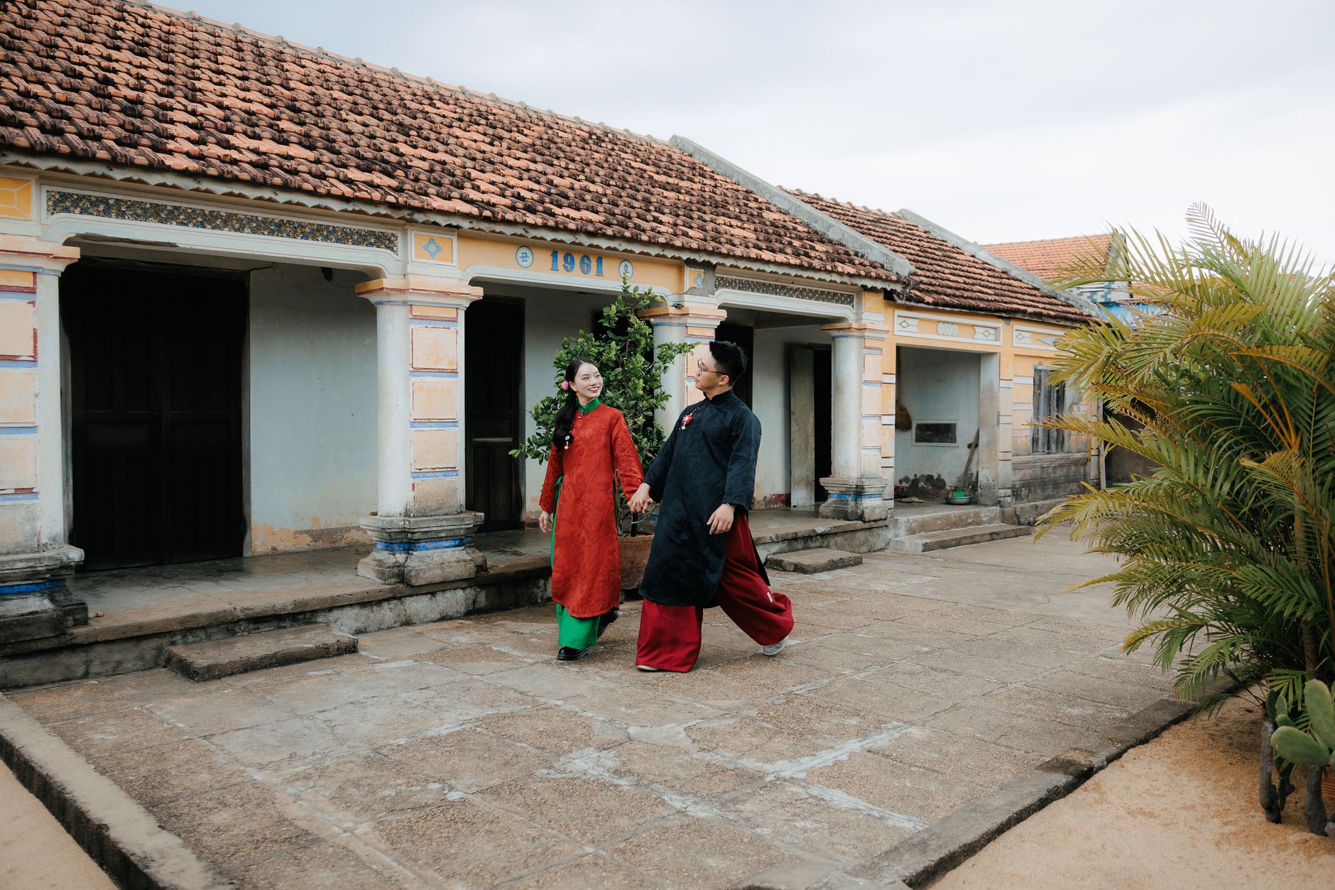 A couple in traditional Vietnamese attire walking by a historical house with 1961 marker.