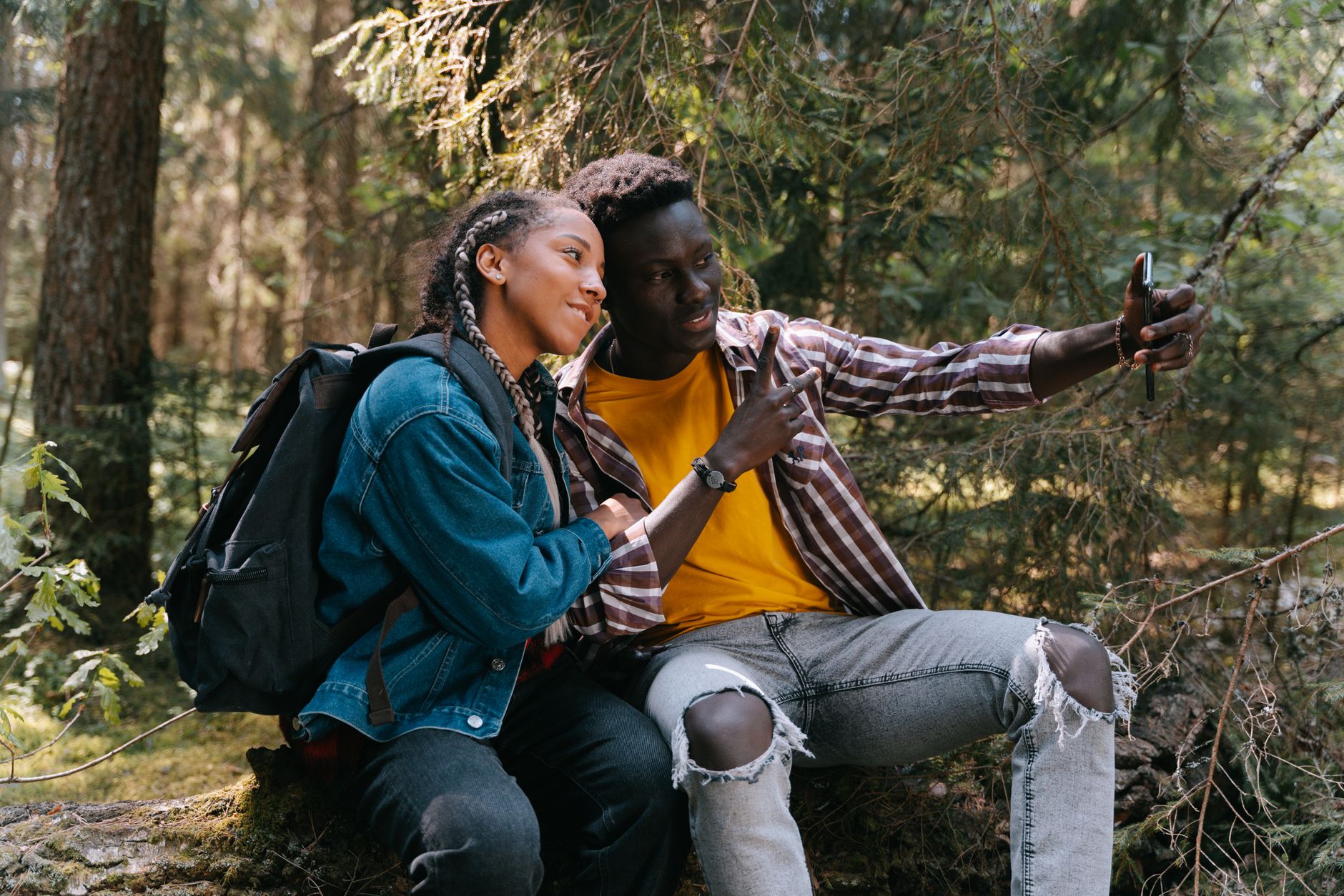 A young couple sits in a forest, capturing a selfie amid nature's tranquility.