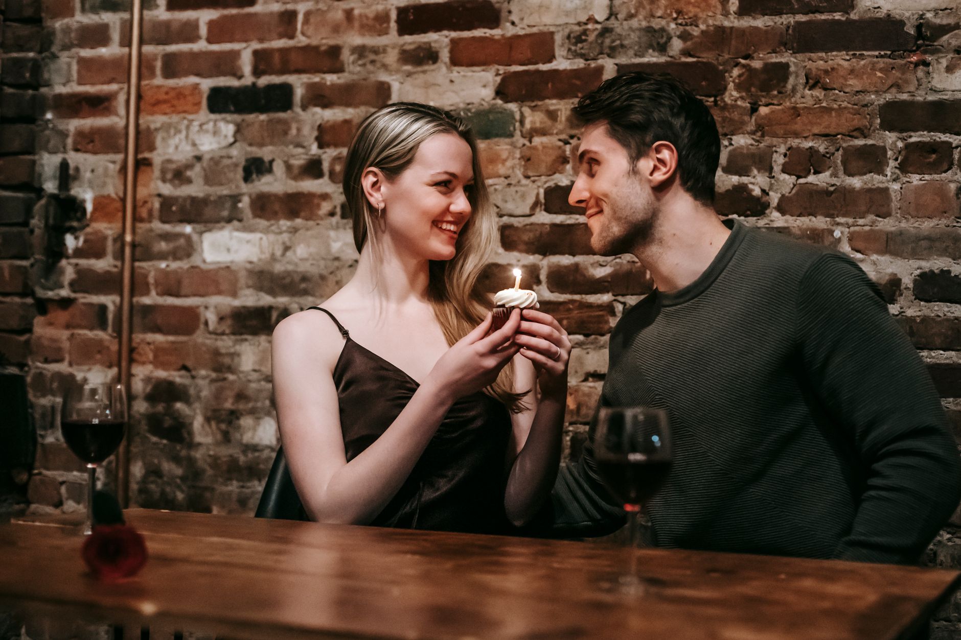 Side view of loving couple sitting near brick wall at wooden table with cupcake with burning candle looking at each other while celebrating…