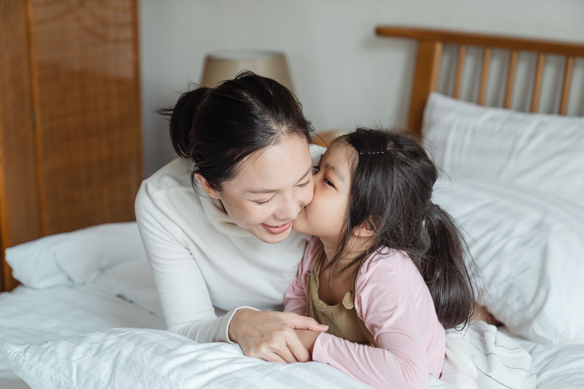 Cute Asian girl kissing on cheek and bonding with smiling young mother while lying in comfort bed in morning and enjoying free time together