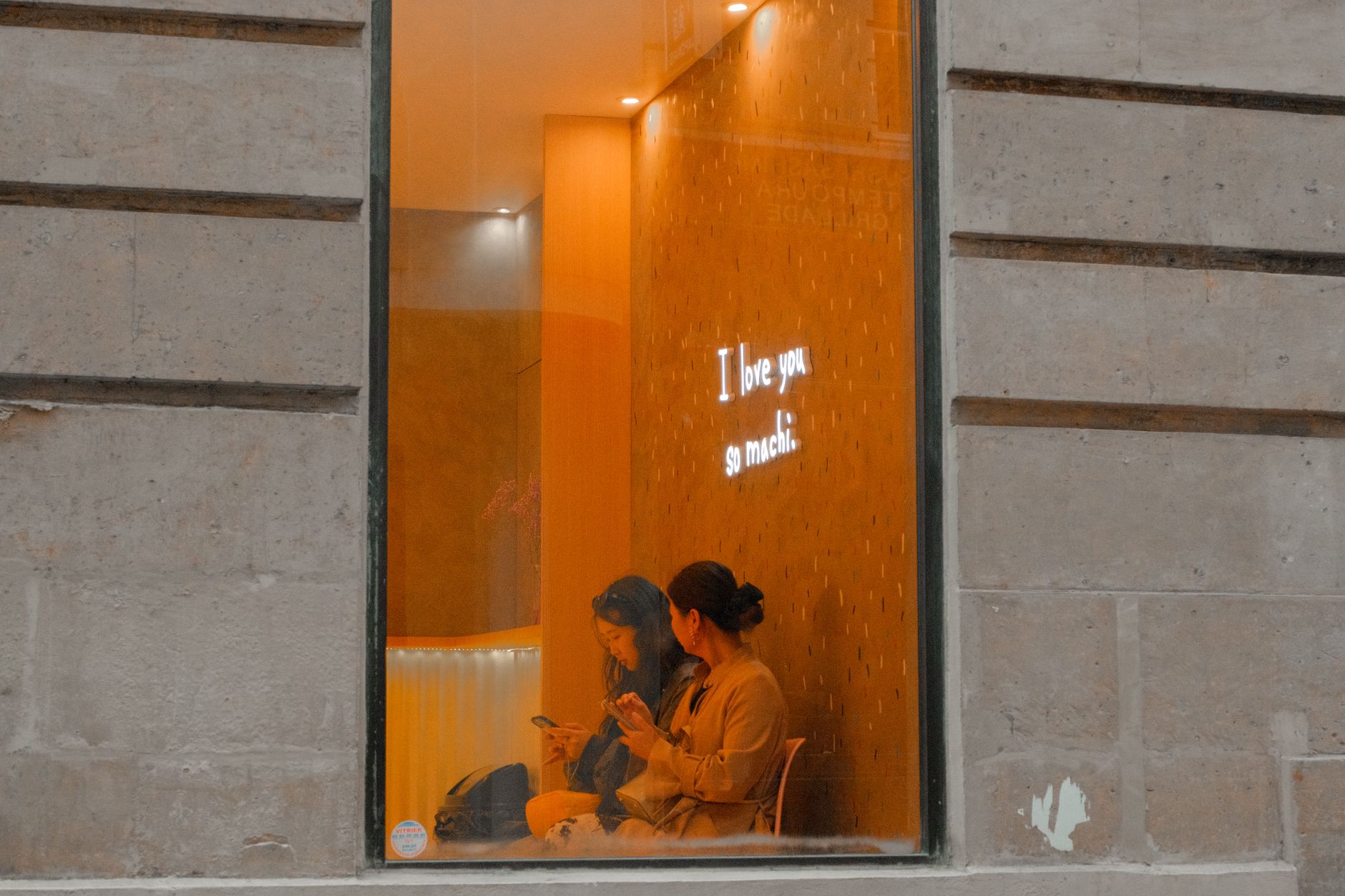 Two women sit in a cozy cafe, viewed through a window with a neon 'I love you so much' sign.