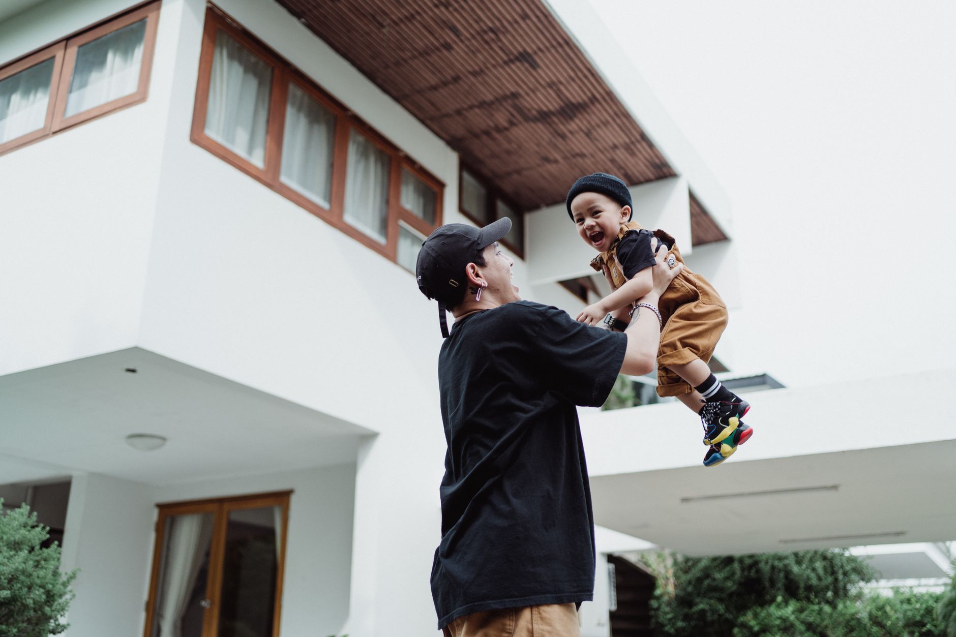 Father joyfully holds his son up in front of a modern house, showcasing happiness and family bonding.