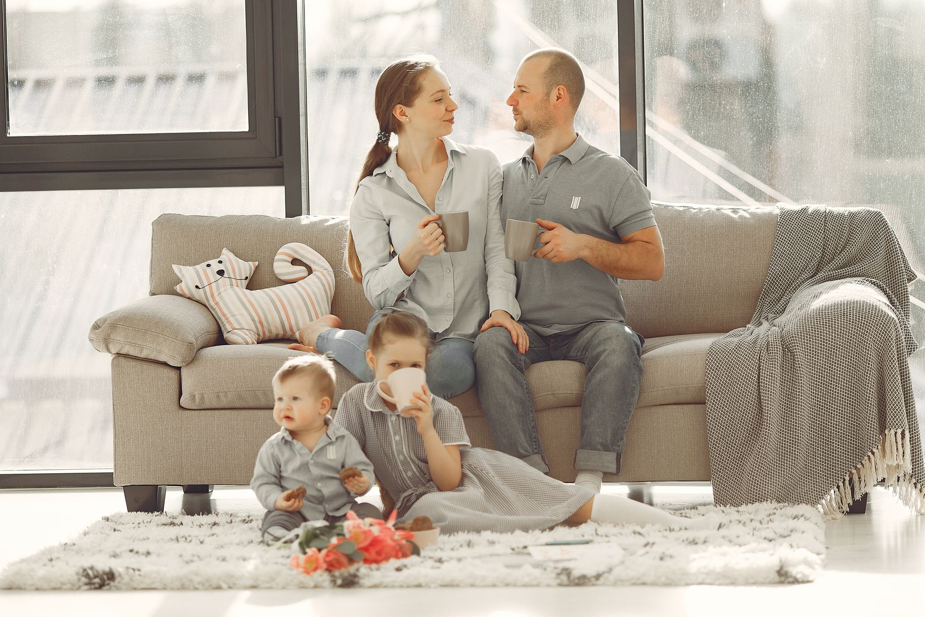 Happy family with children spending time at home while parents sitting on couch with cups of coffee and looking at each other when kids res…