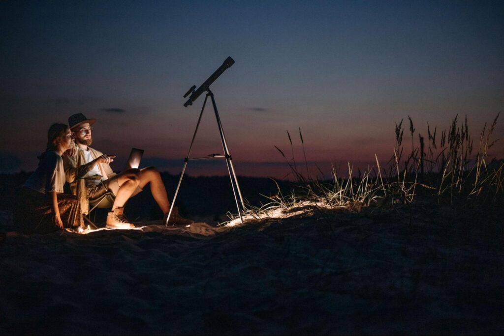 Couple stargazing together with a telescope on the sand at night