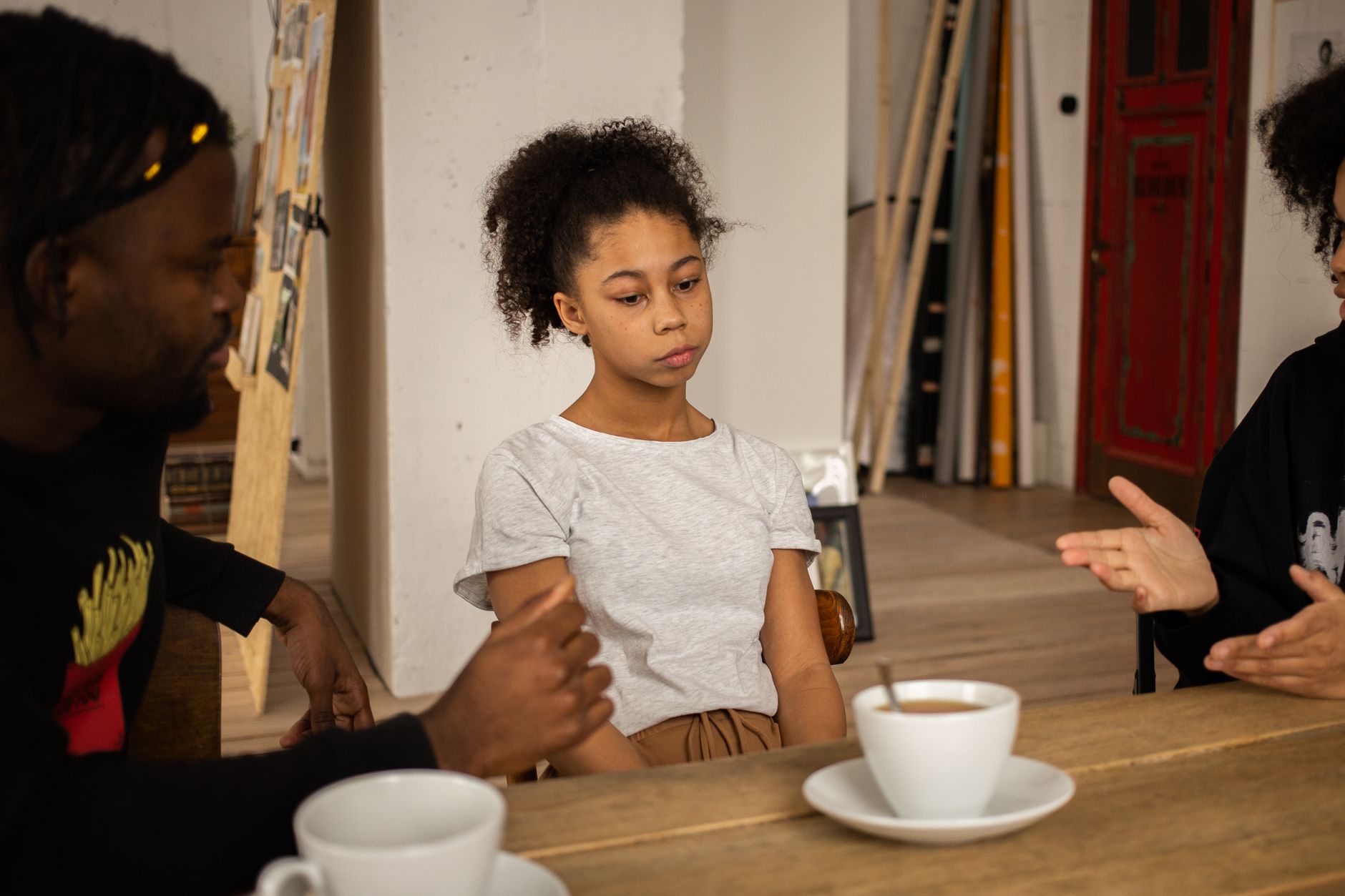 Sad African American girl sitting at wooden table with cups of coffee near black parents having conversation in light room