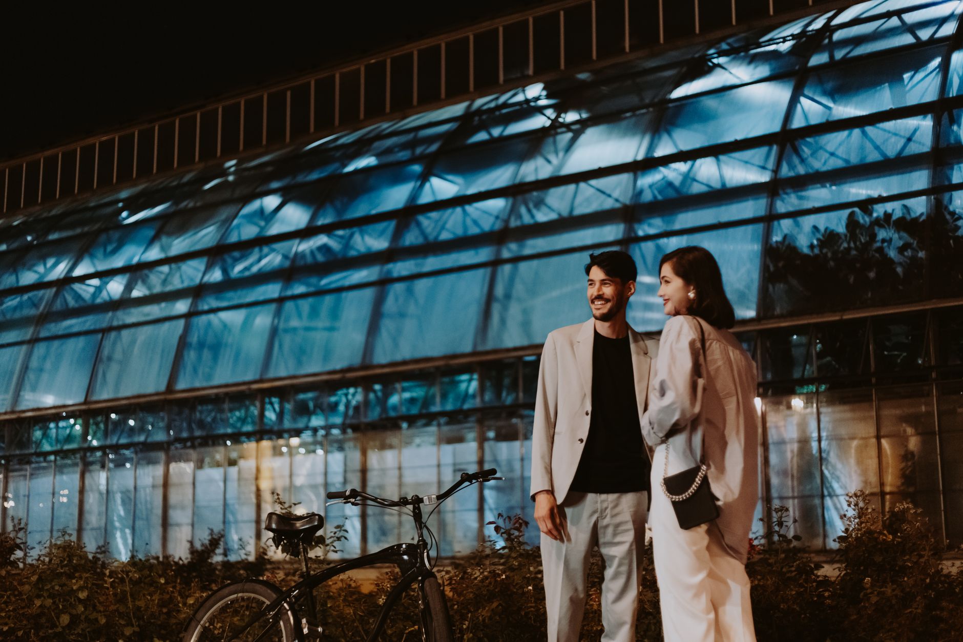 Couple enjoying a night out by a modern glass building with a bicycle.