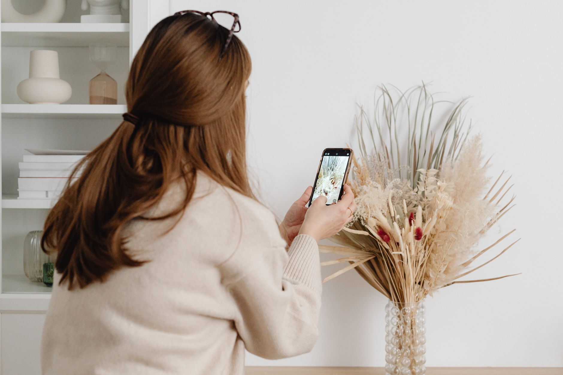 A woman takes a picture of a dried floral arrangement in a stylish home setting.
