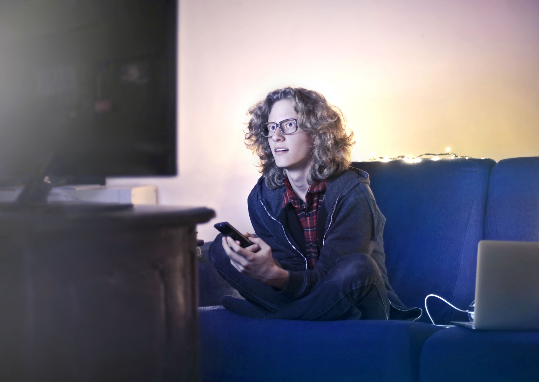 Concentrated male with long hair sitting on comfortable sofa at home and messaging on social media via cellphone while watching movie on TV…